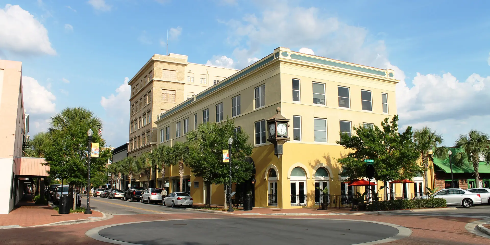 A pale yellow three-story corner building with a street clock in downtown Winter Haven, palms along a brick sidewalk